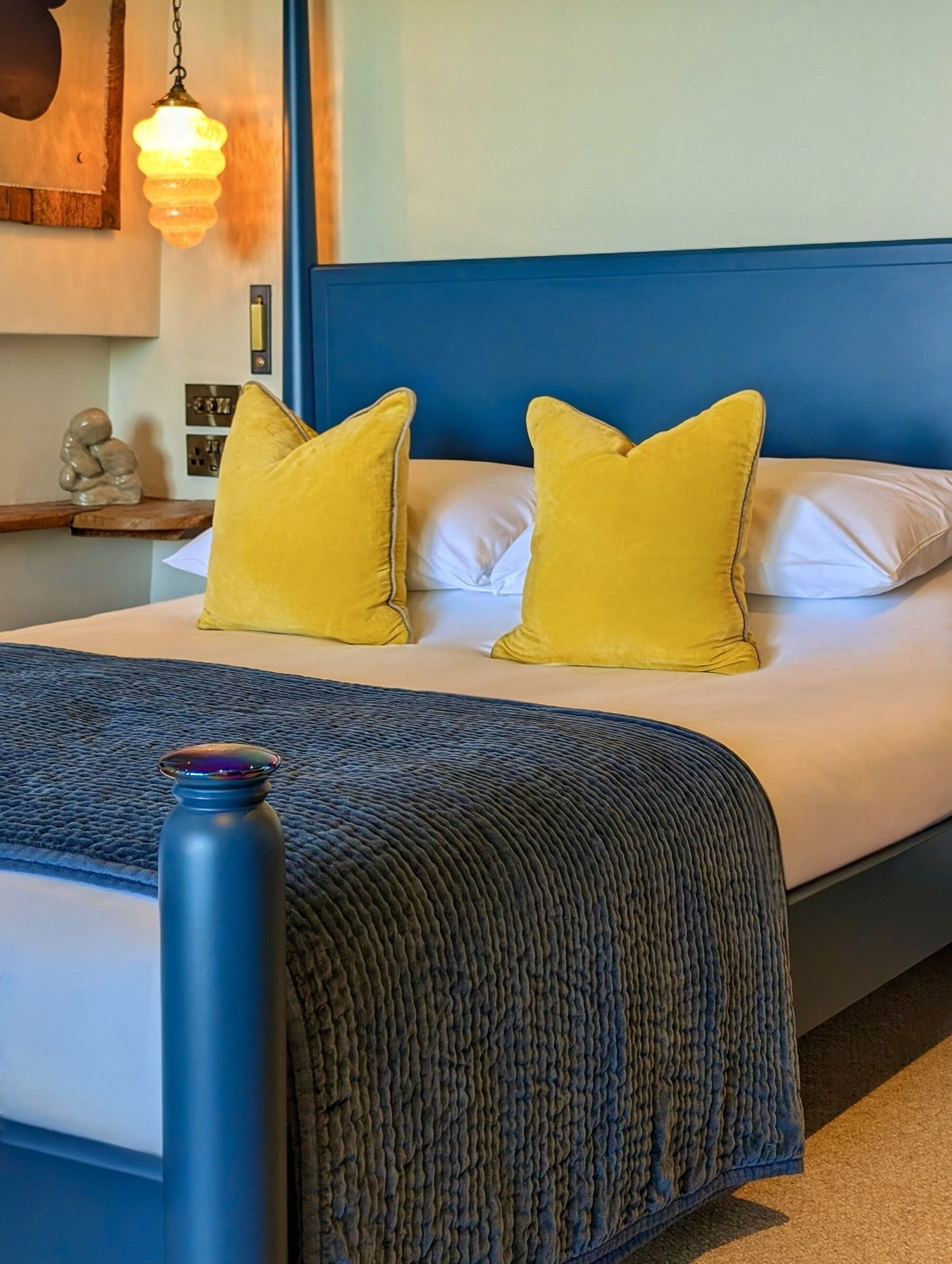 A bed with a blue headboard, two yellow pillows, a white pillow, and a dark blue textured blanket.