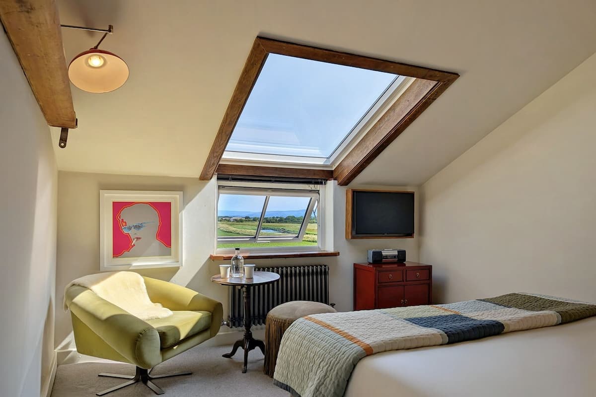 A bedroom with a skylight, a bed, a chair, a small table with a bottle, a radiator, and a TV.