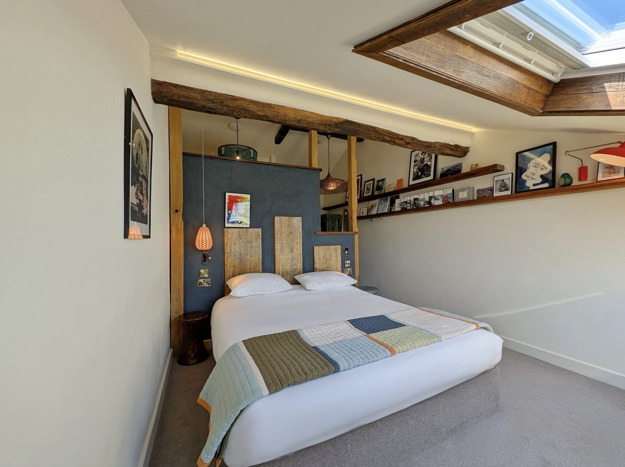 Un dormitorio con una cama grande, tragaluz, techo con vigas de madera y estante con cuadros enmarcados.