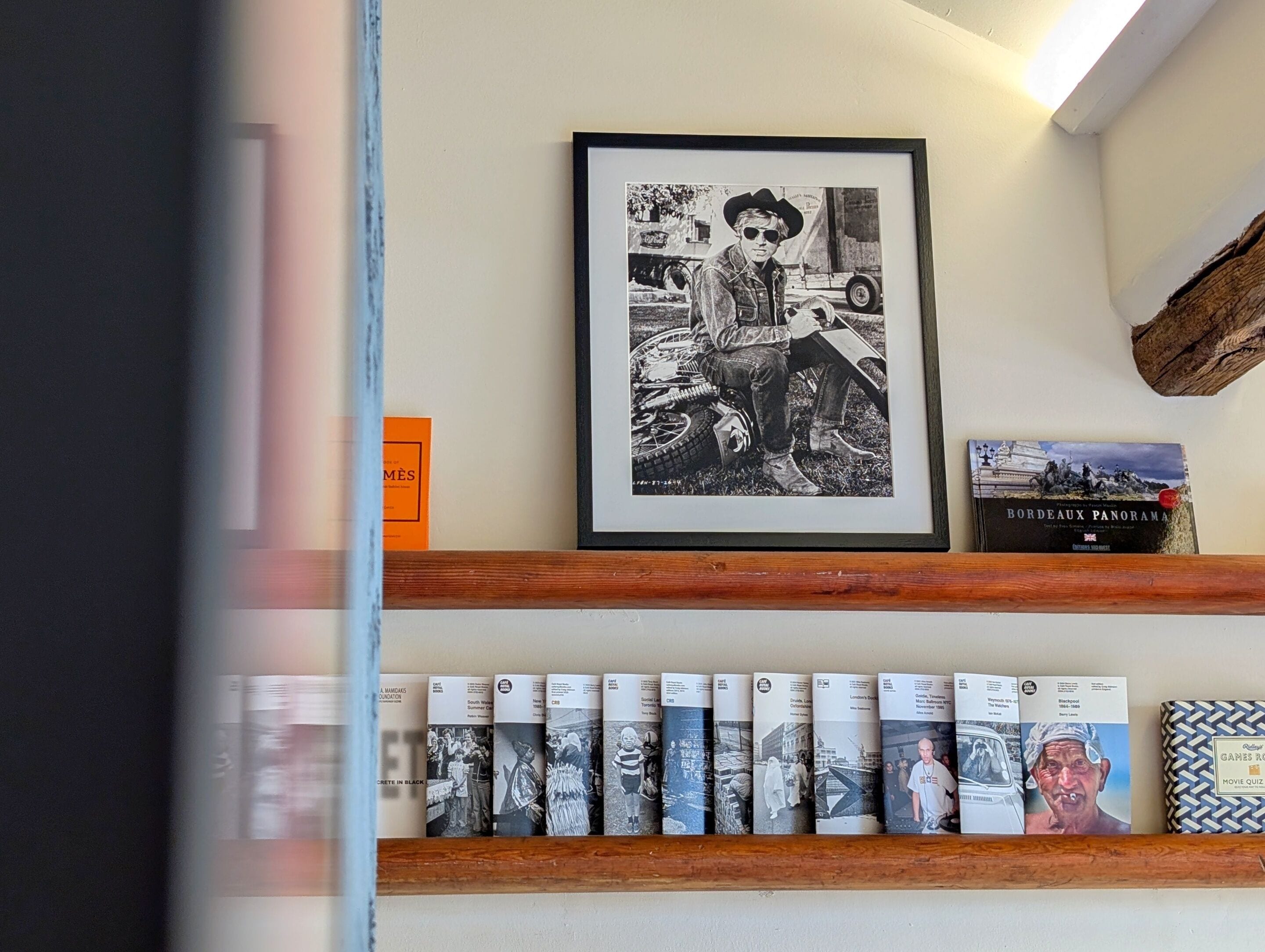 Ein gerahmtes Foto eines Mannes mit einem Motorrad an einer Wand, ein Bücherregal mit Büchern und ein gerahmtes Buch mit dem Titel 'Bordeaux Panorama'.
