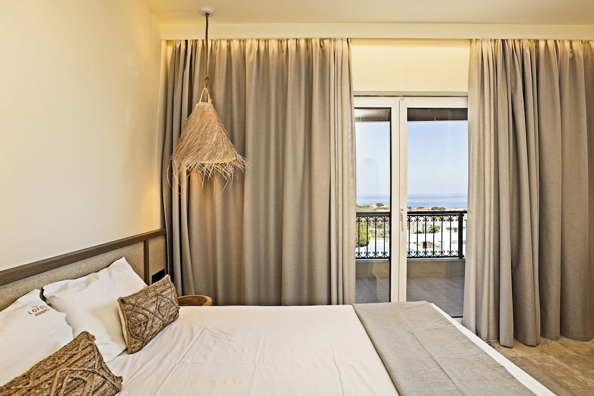Habitación de hotel con cama, cortinas, lámpara colgante y vista a un balcón con barandilla.