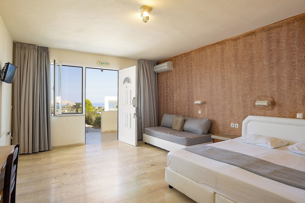 Una habitación de hotel con cama, sofá, televisión y puerta corredera que da a un balcón.