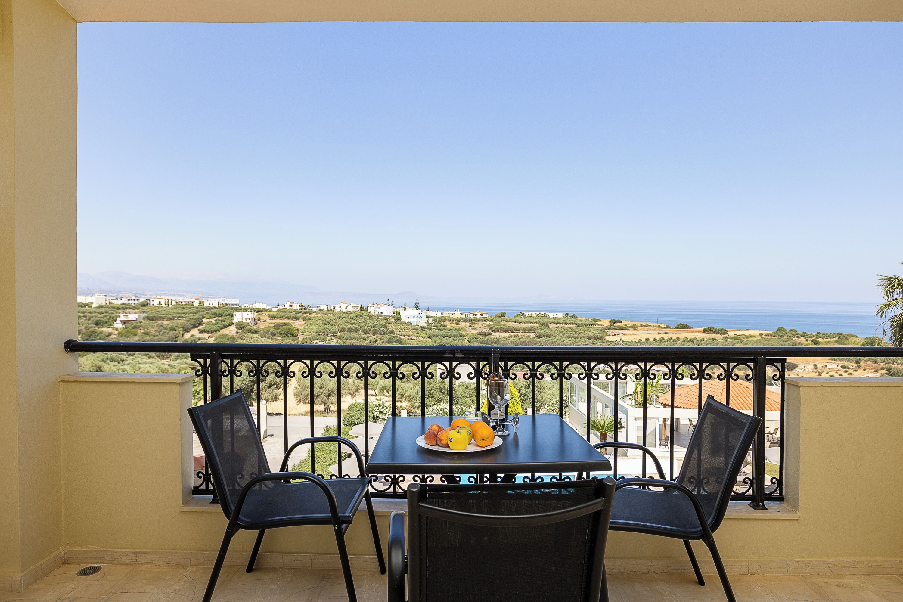 Un balcon avec une table et des chaises, une assiette de fruits et une vue sur la mer et les collines.