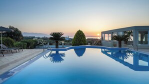 Une piscine à l’eau d’un bleu cristallin, entourée d’un bâtiment moderne doté de grandes fenêtres et offrant une vue sur le coucher de soleil.