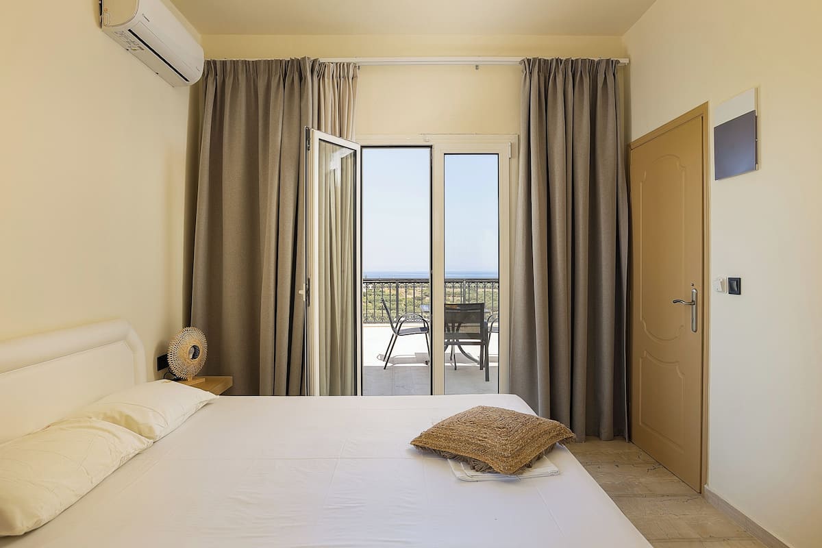 Una habitación de hotel con una cama, una mesita de noche con lámpara, una ventana con cortinas y una puerta que da a un balcón.