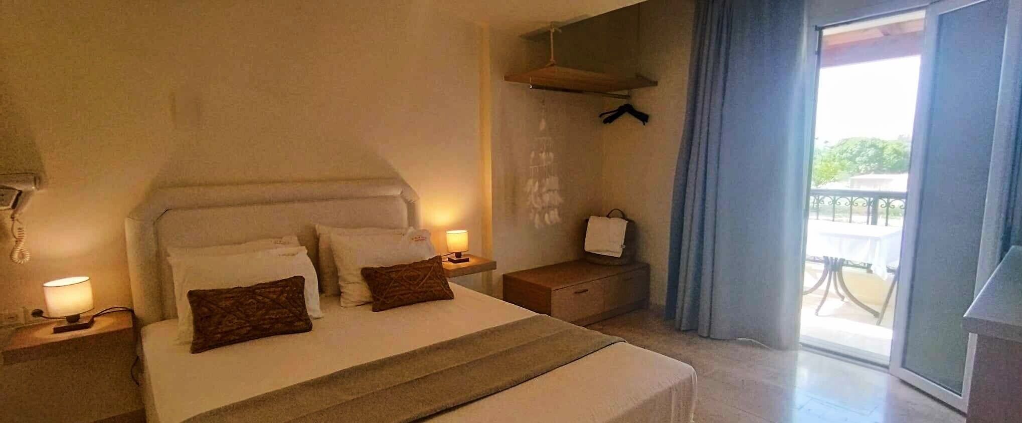 A hotel room with a bed, bedside lamps, a small table, a hanging towel, and a view of the outdoors.
