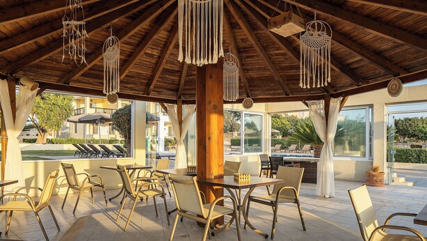 Un espace extérieur aménagé avec des tables et des chaises en bois, un grand parasol et des décorations en macramé suspendues.