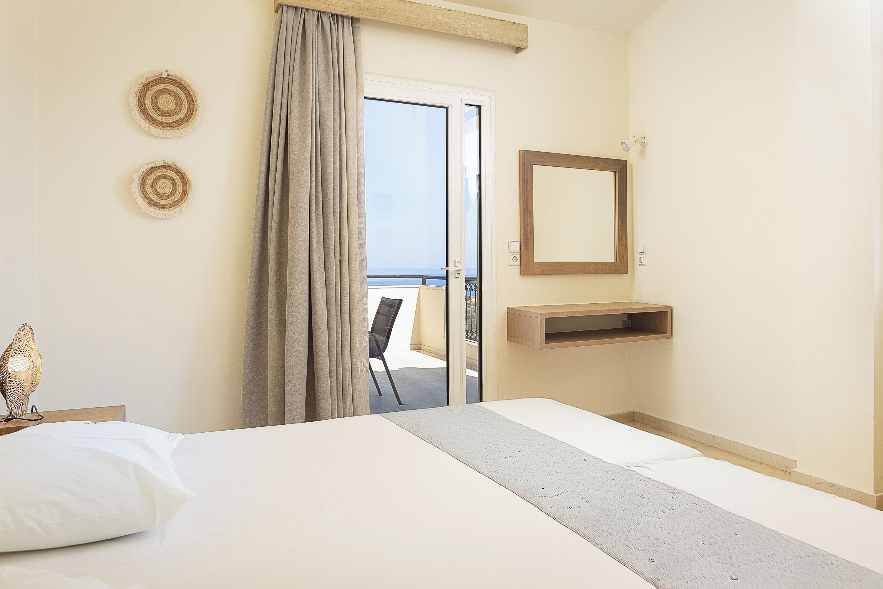 A hotel room with a bed, a bedside table with a lamp, a mirror, a small shelf, and a view of the balcony.
