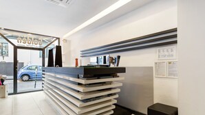 A reception desk with a glass door entrance, a blue car parked outside, and a black trash bin to the right. - Golden Hotel (Naples)