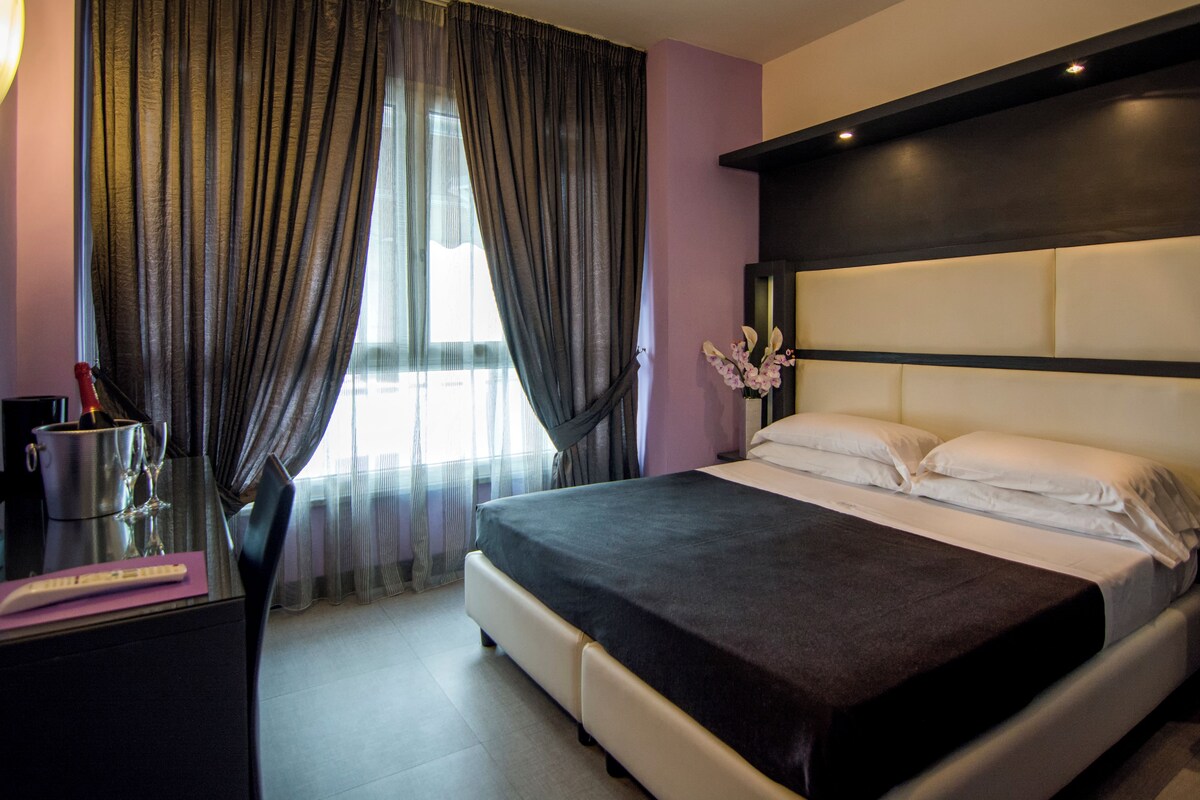 A modern hotel room with a large bed, a desk with a chair, and a vase of flowers.