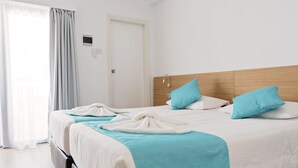 Une chambre d’hôtel avec une tête de lit en bois, deux lits avec une literie blanche et turquoise, une porte blanche et une fenêtre avec des rideaux.