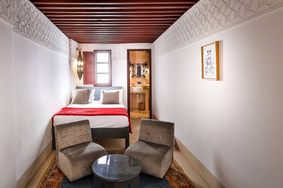 A narrow room with a bed, two armchairs, a small table, and a door leading to a bathroom.