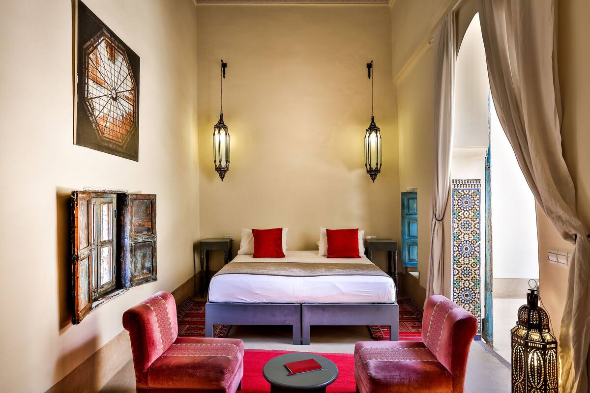 A hotel room with a large bed, two red chairs, a small table, and a decorative lantern.