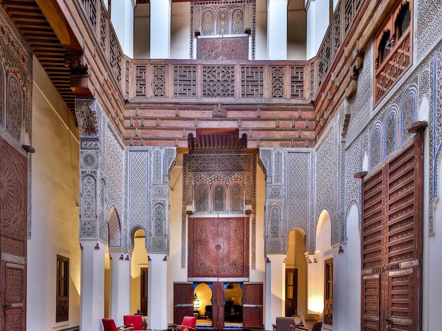 A courtyard with intricate tile work, a central seating area with red chairs, and a wooden ceiling.