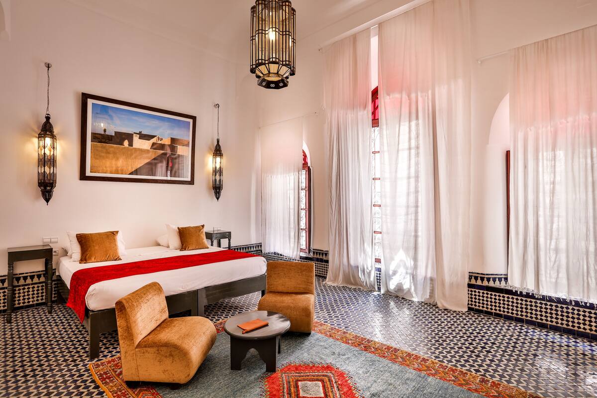 A hotel room with a bed, two chairs, a small table, a patterned rug, and a large window with sheer curtains.