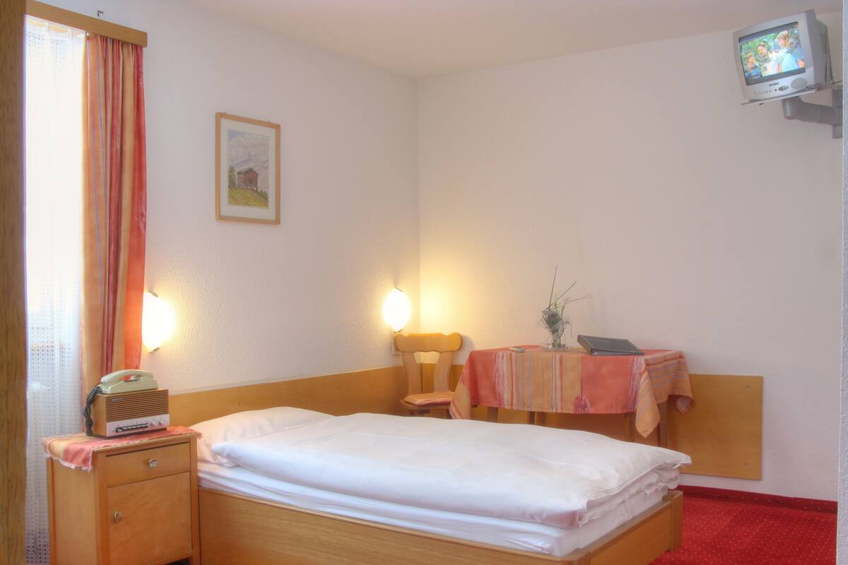A hotel room with a single bed, bedside table, lamp, chair, small table with a vase, and a television on a stand.