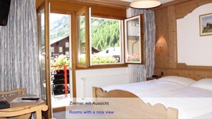 A bedroom with a bed, a chair, and a window with a view of a mountain and a building. - Hotel Restaurant Mattmarkblick (Saas Almagell)