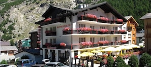 A hotel with a terrace and outdoor seating area, surrounded by parked cars and a mountainous backdrop. - Hotel Restaurant Mattmarkblick (Saas Almagell)