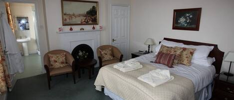 A bedroom with a large bed, two chairs, a nightstand, a lamp, and a painting on the wall.
