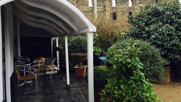 A covered outdoor seating area with chairs and tables, a stone wall, and a large bush. - Castle House Hotel (Canterbury)