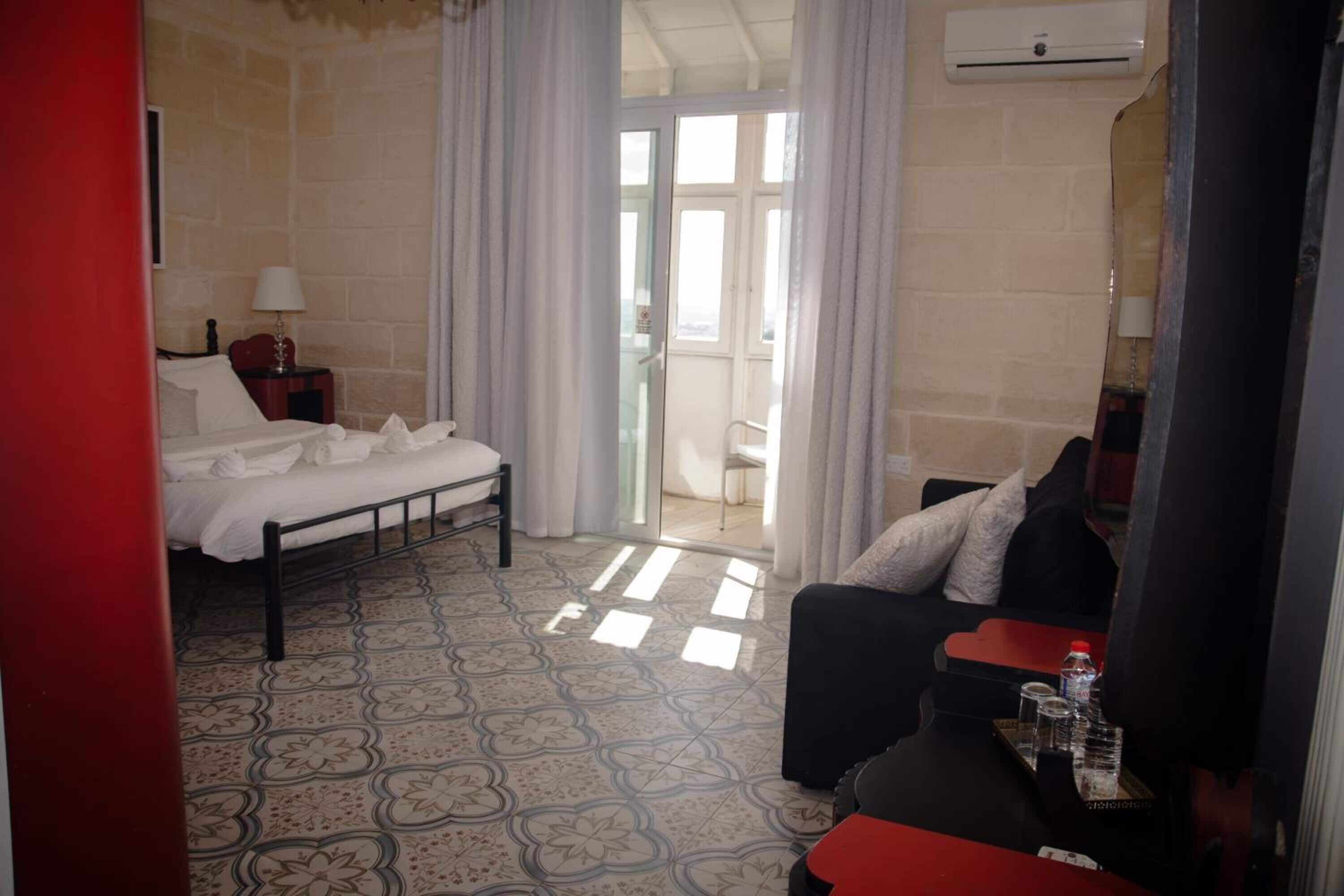 A hotel room with a bed, a sofa, a small table with a bottle and glasses, and a window with curtains.