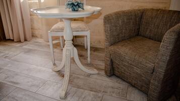 A white table with a decorative blue flower centerpiece, a white chair with a cushion, and a brown woven sofa in a room with stone walls and tiled flooring.