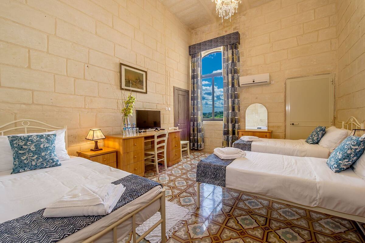 A hotel room with two beds, a desk, a TV, and a view of the sky through a window.