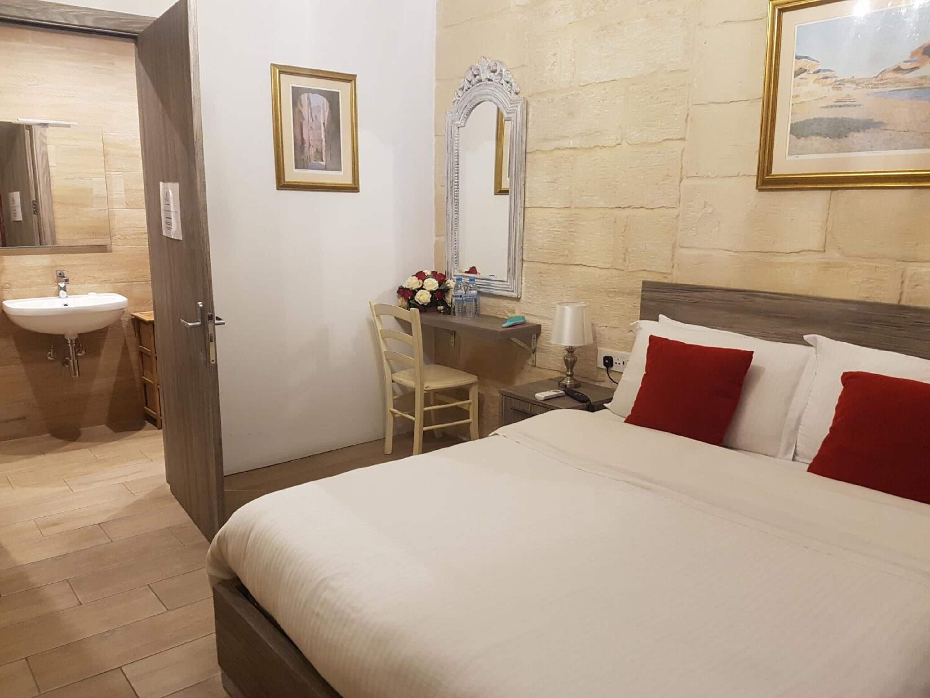 Habitación de hotel con cama, escritorio con silla, espejo, lavamanos y un cuadro en la pared.