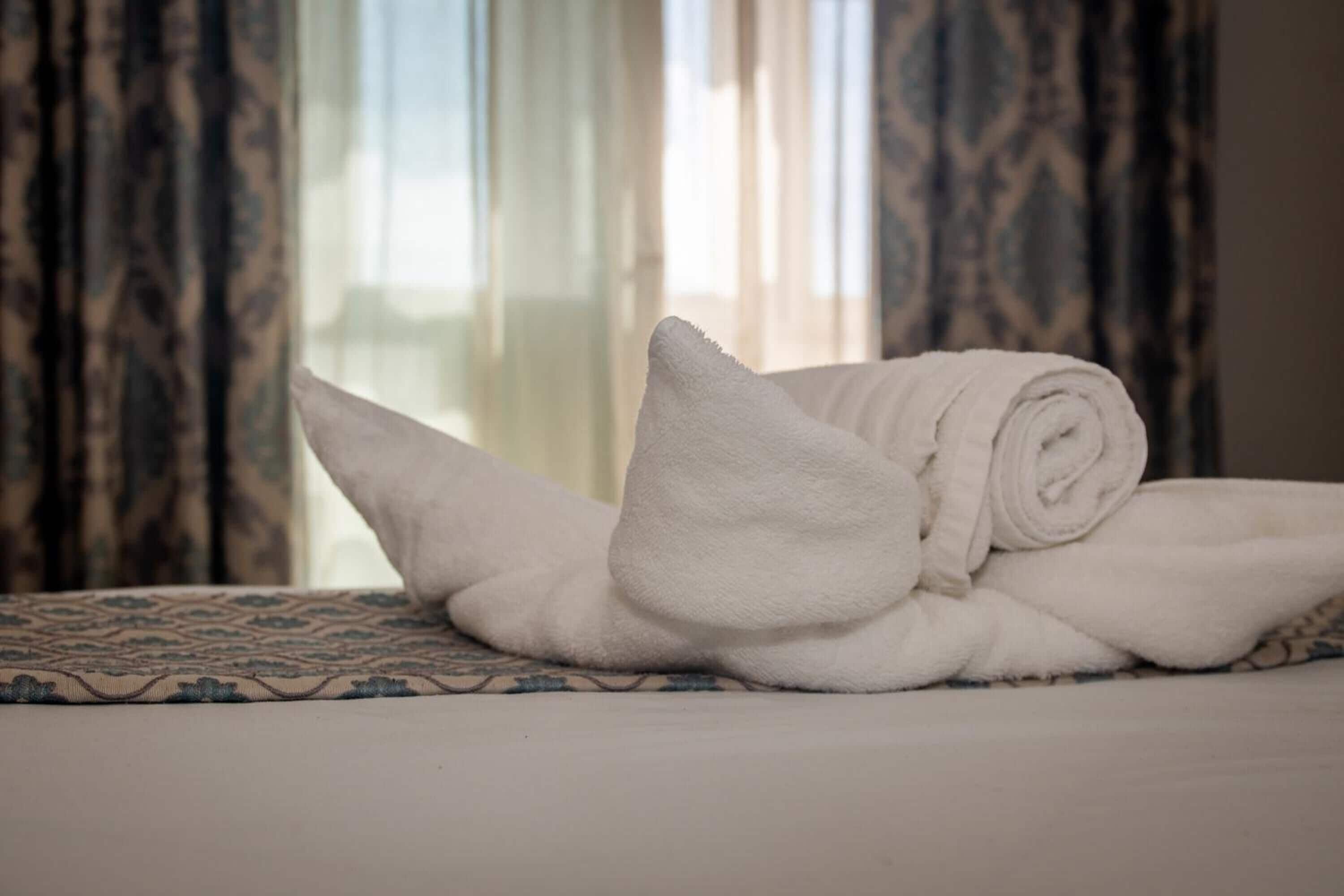 Una cama bien hecha con una toalla enrollada y un almohadón sobre una colcha estampada.