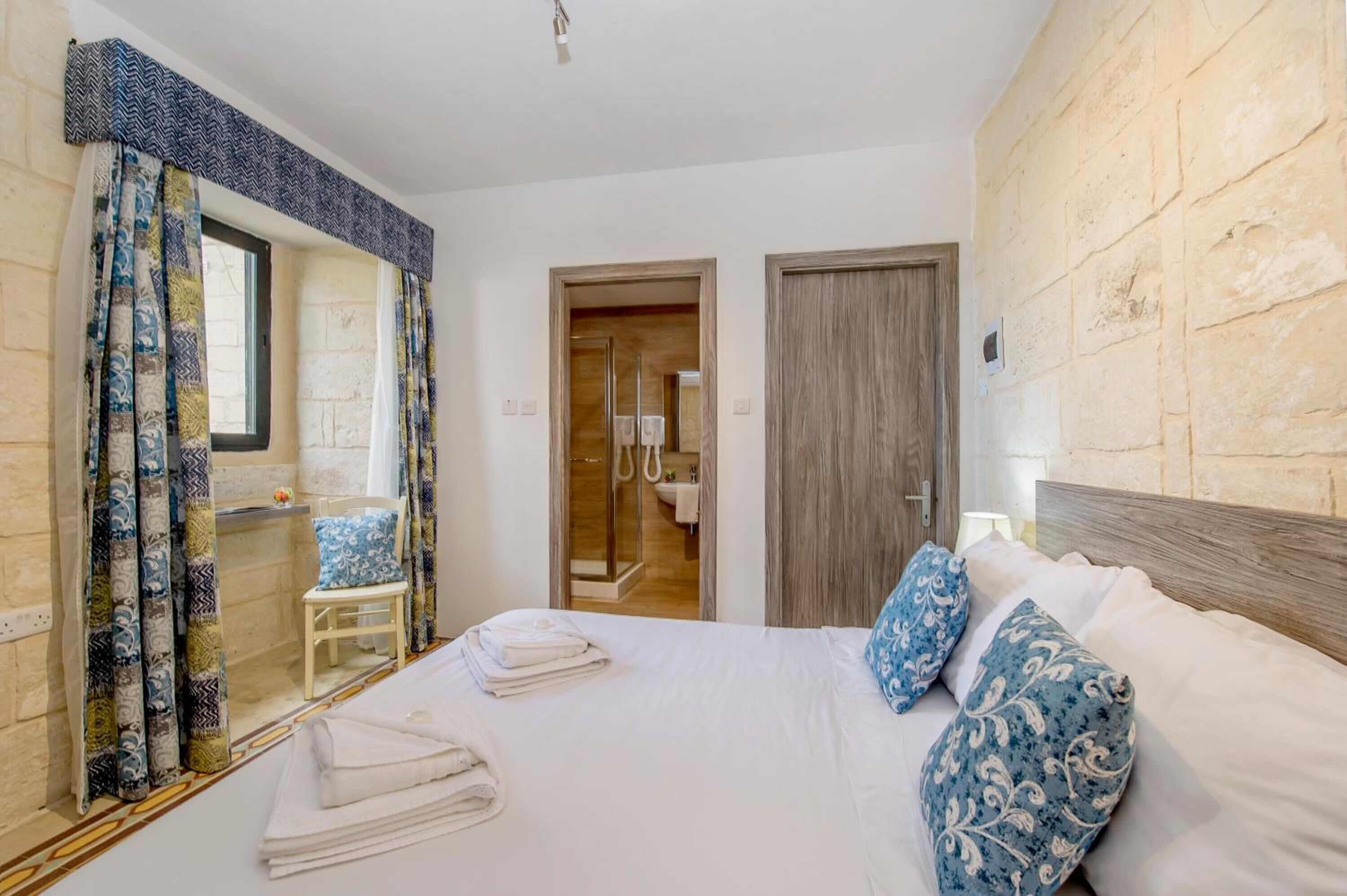 Una camera d'albergo con un letto grande, una testiera in legno, uno specchio, una finestra con tende a motivi e una porta che dà su un bagno.
