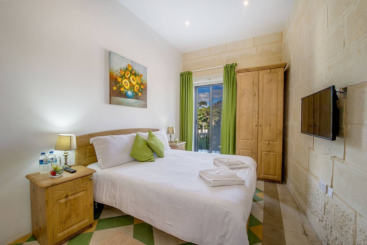 A bedroom with a bed, bedside tables, a painting, a TV, and a view through a window with curtains.