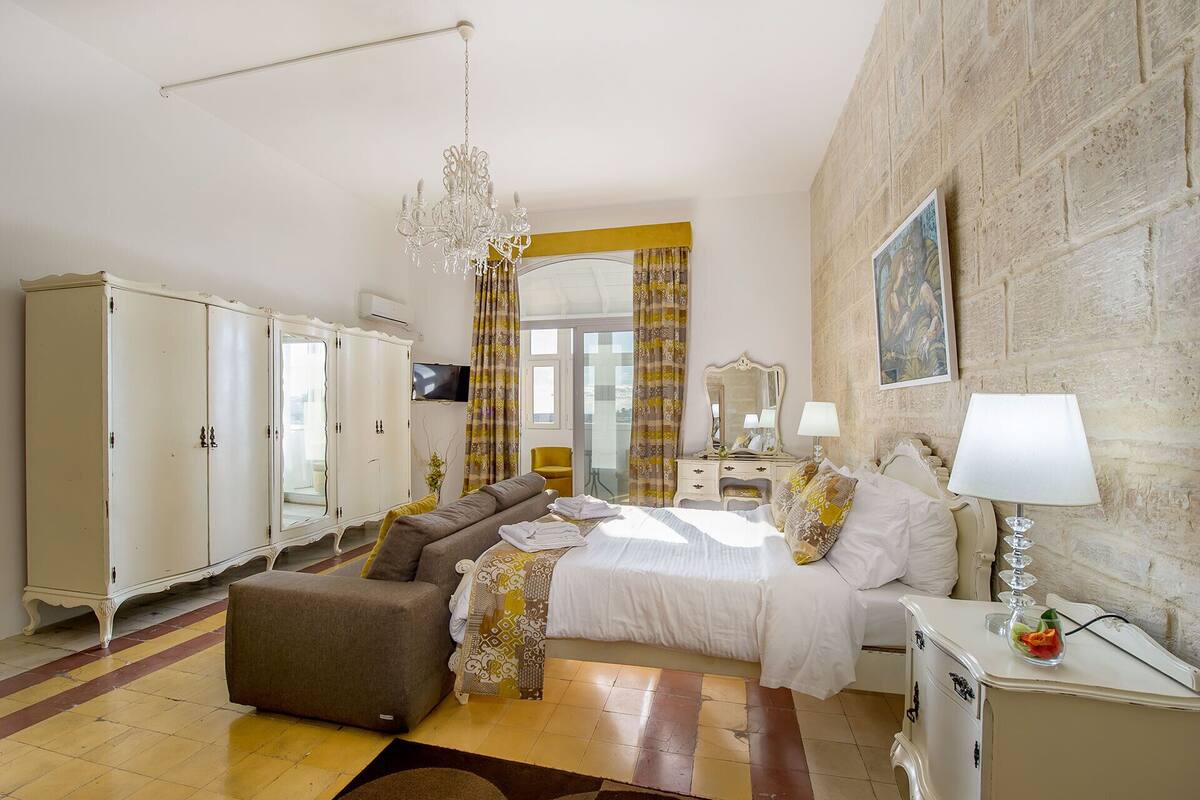 A hotel room with a bed, a sofa, a chest of drawers, a television, and a stone wall.