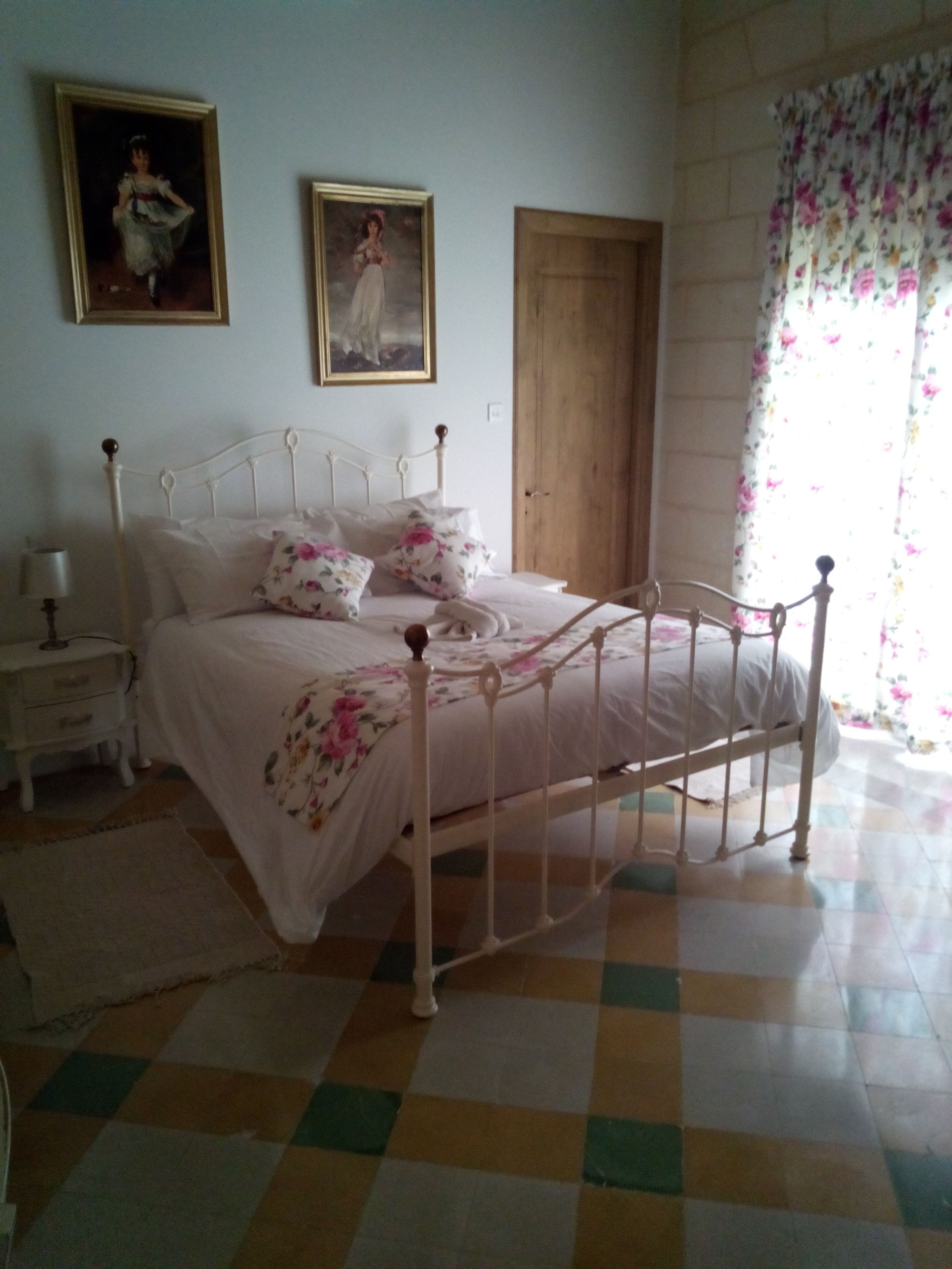 A bedroom with a checkered floor, a bed with floral bedding, a nightstand, a lamp, and two framed pictures on the wall.