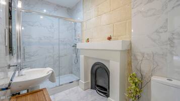A bathroom with a glass shower enclosure, a white sink, a marble-tiled floor, a fireplace, and a toilet.