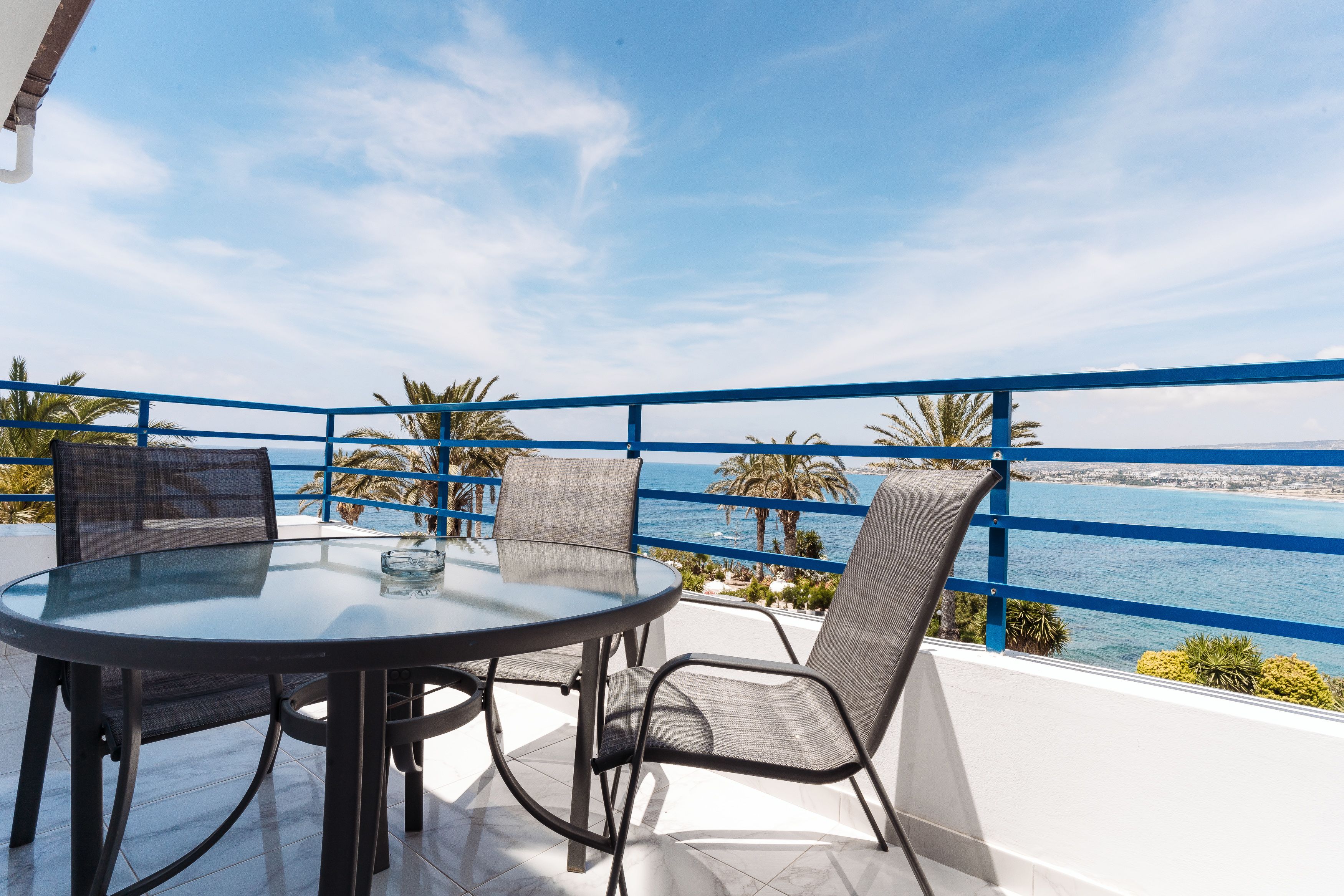 A round glass table with metal chairs on a balcony with a blue railing, overlooking a coastal area with palm trees and a view of the sea.