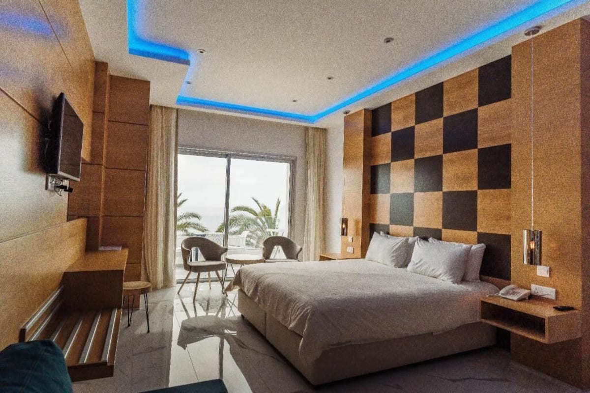 A modern hotel room with a large bed, a TV mounted on the wall, a balcony with chairs, and a view of palm trees.