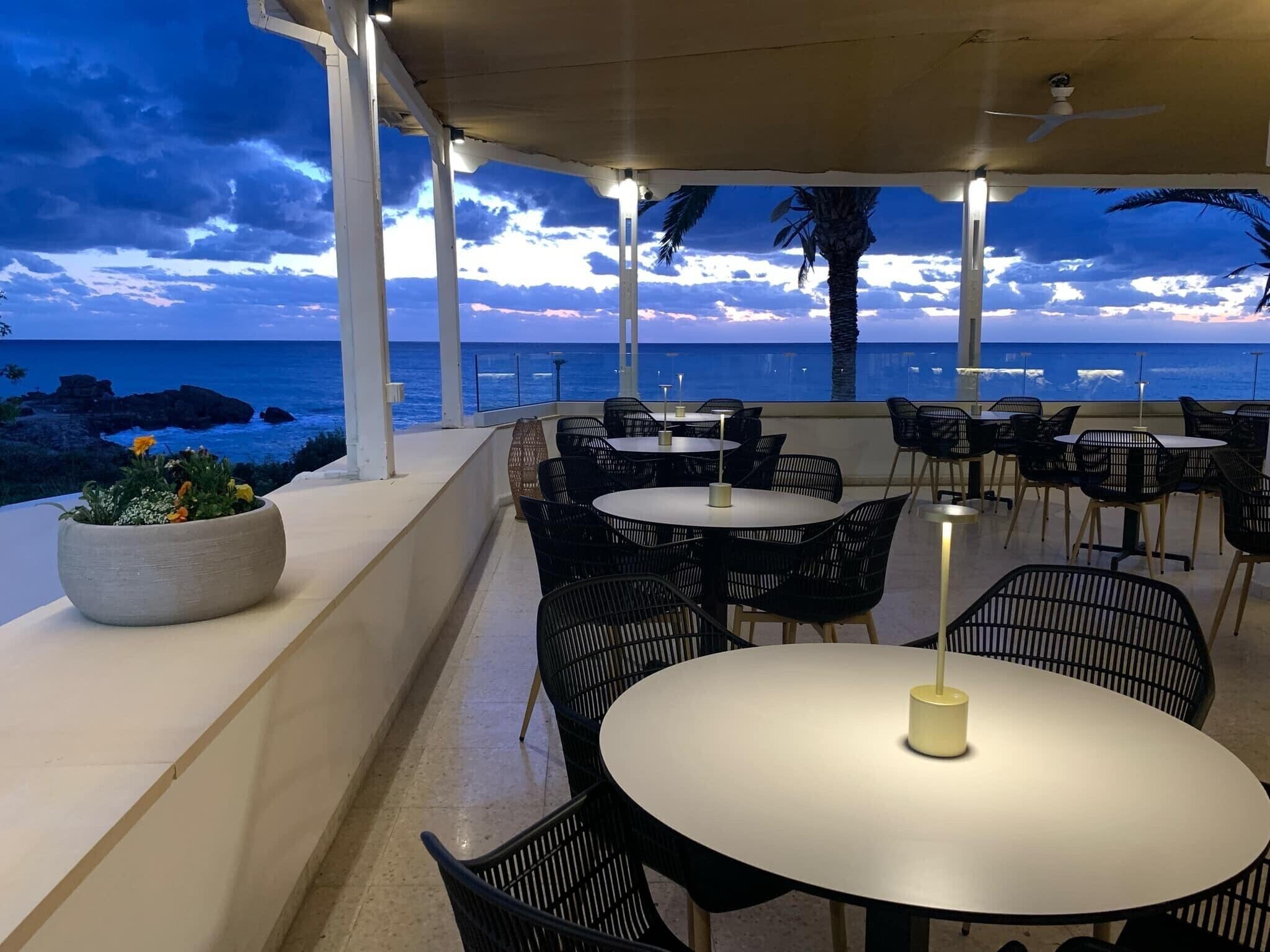An outdoor patio with round tables and chairs, overlooking the ocean with a view of the sea and rocks.