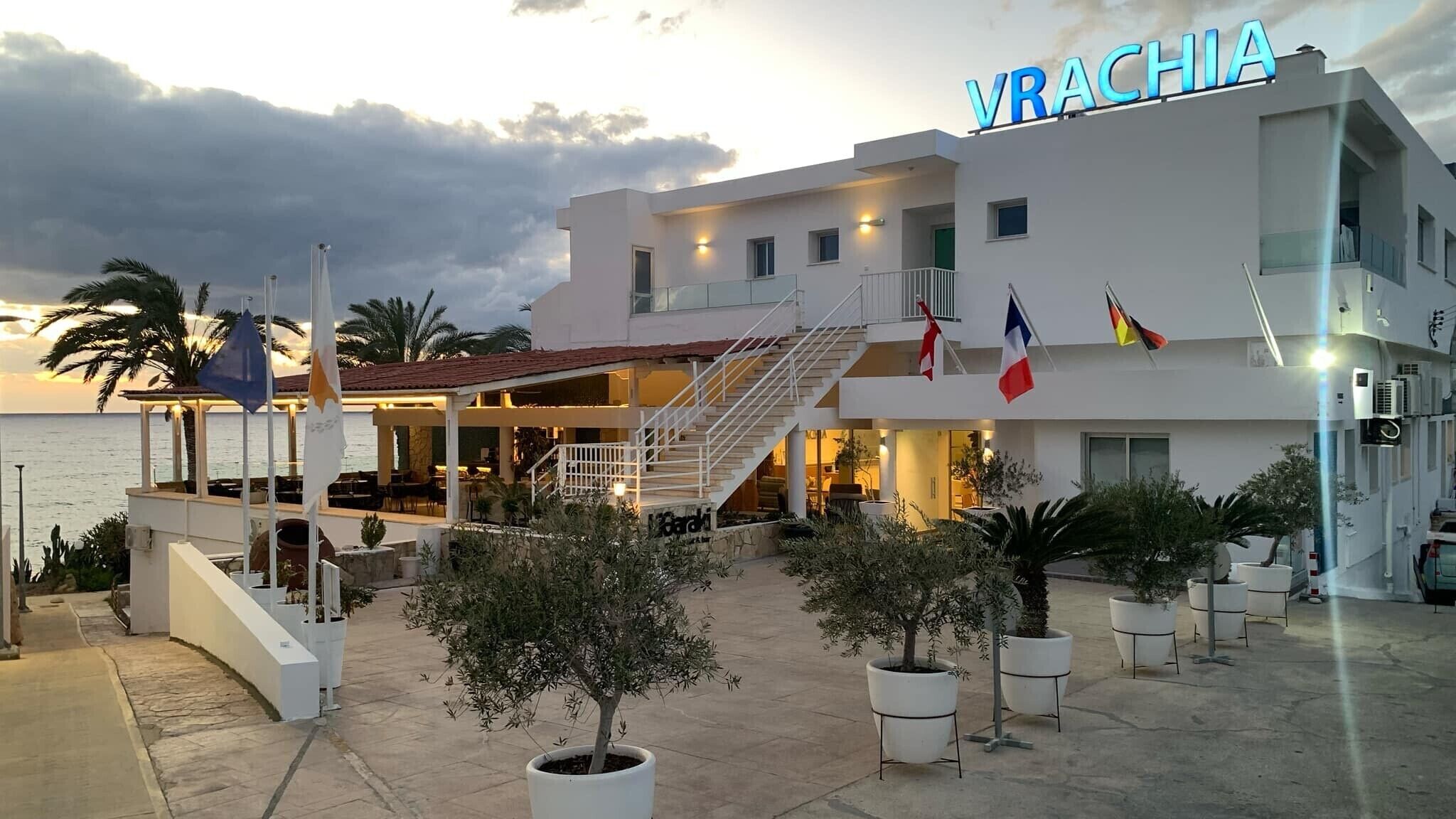 Un bâtiment moderne avec une enseigne indiquant « VRACHIA » et plusieurs drapeaux. On y trouve un espace extérieur abrité avec des tables et des chaises, ainsi que des plantes en pot tout autour.