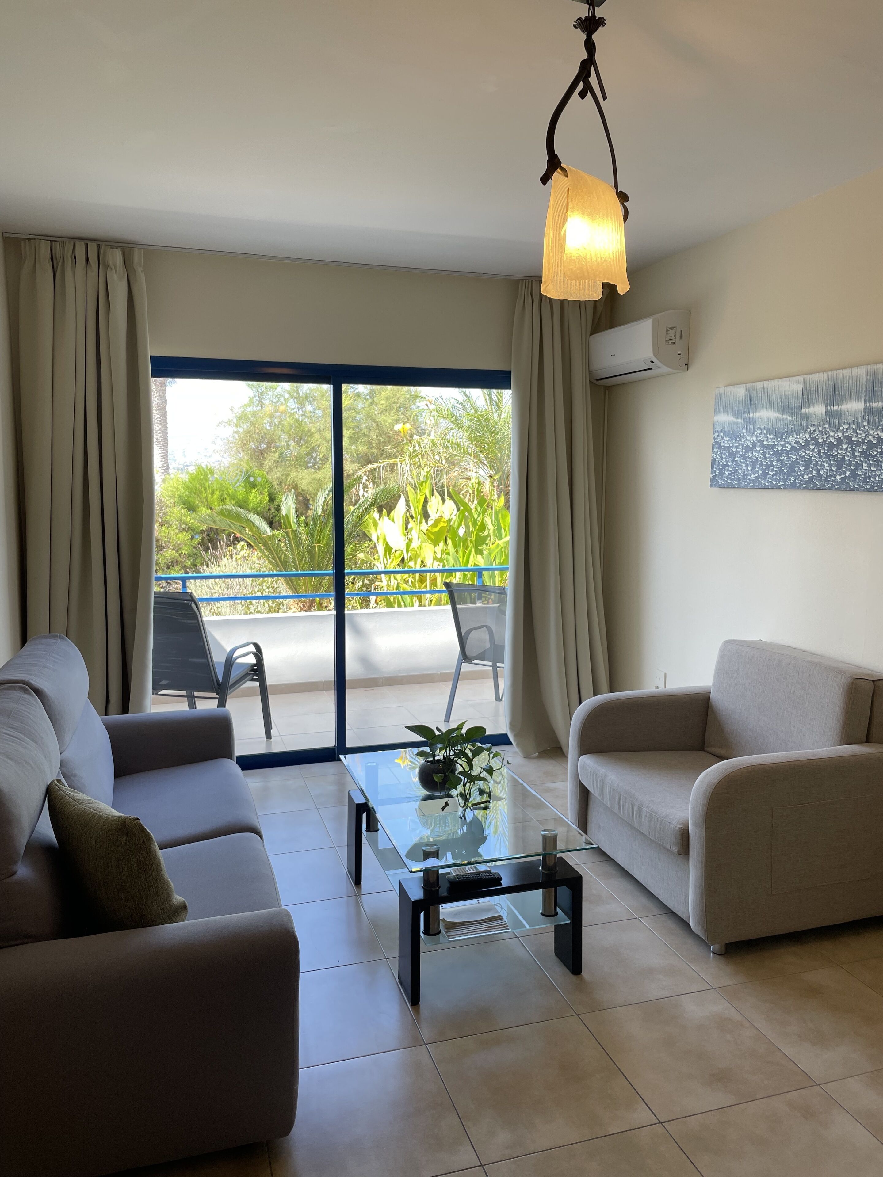 A modern living room with two sofas, a glass coffee table, and a ceiling light. A sliding glass door leads to a balcony with a view of greenery.