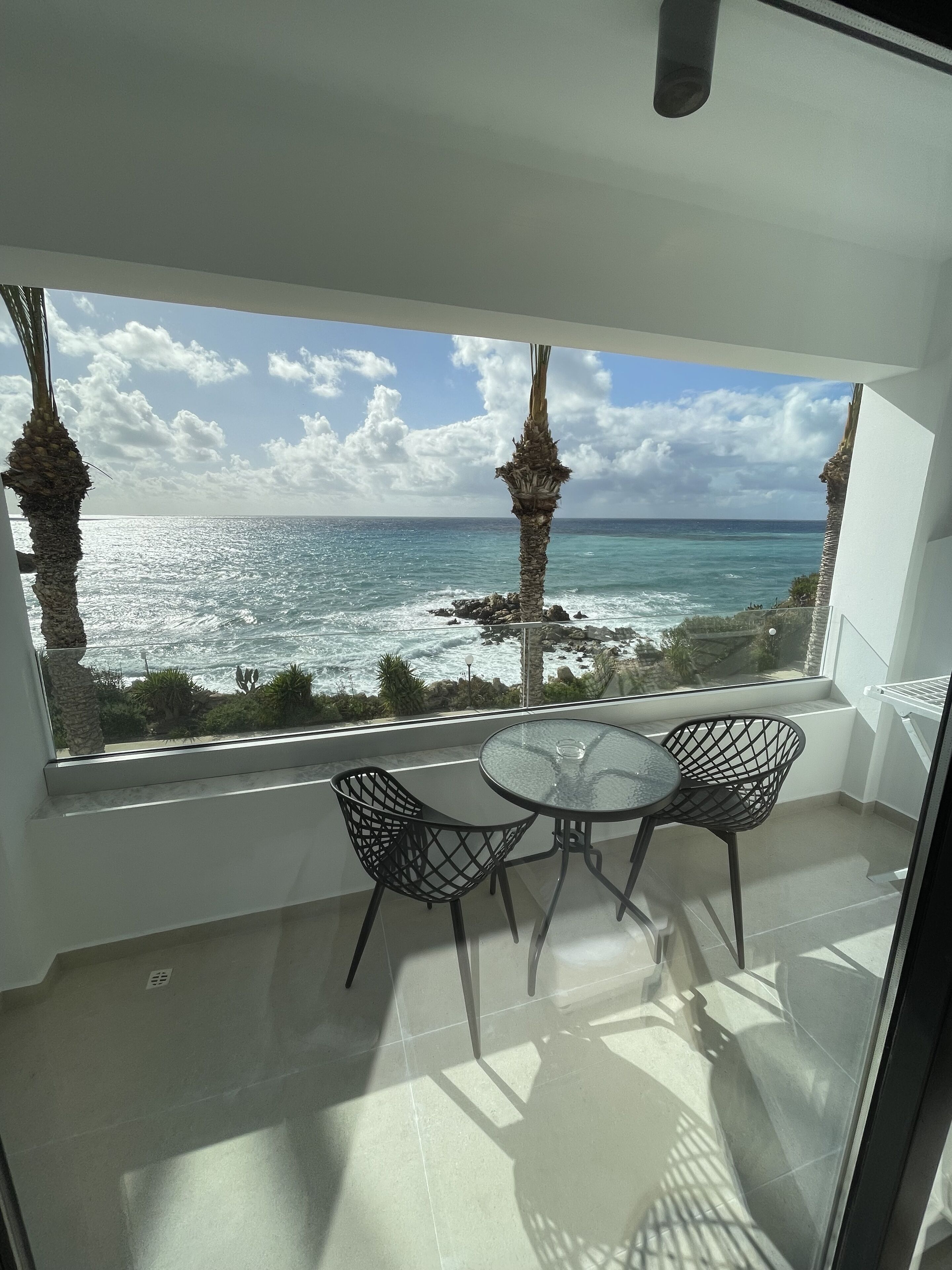 Un balcón con una mesa y sillas de vidrio, con vistas a la costa, palmeras y el océano.