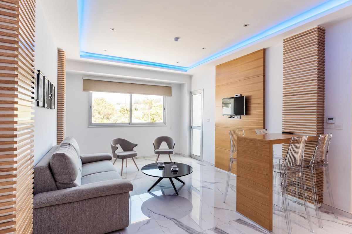 A modern living room with a sofa, two chairs, a coffee table, a wooden accent wall, a TV mounted on the wall, and a large window with a view of trees.