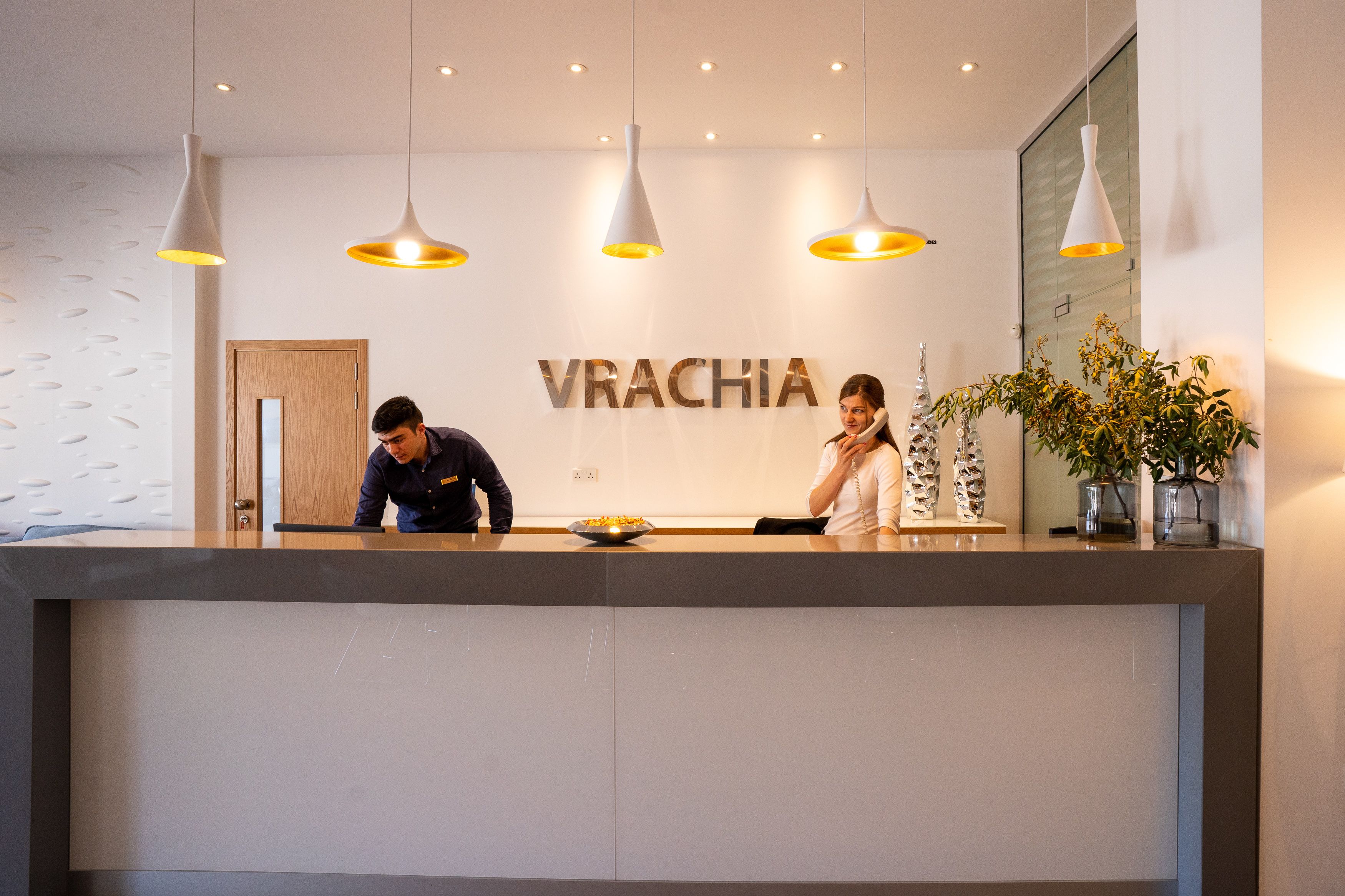 A reception desk with the name 'VRACHIA' prominently displayed. There are three pendant lights above the desk, and a potted plant is visible to the right.