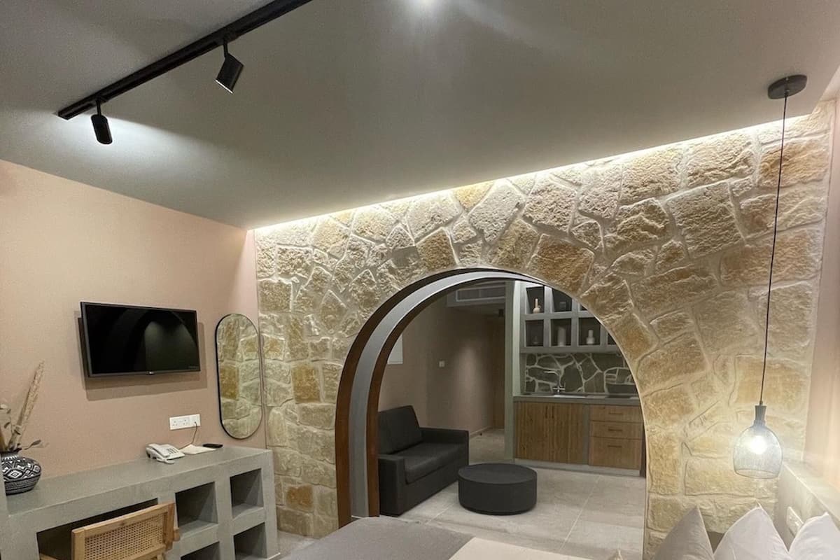 A modern bedroom with a stone archway leading to a living area, a bed with pillows, a desk with a TV, and a hanging light fixture.