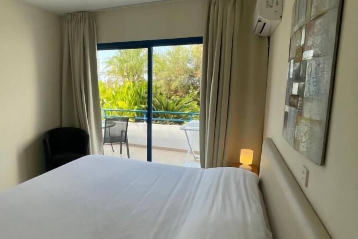 A hotel room with a bed, a chair, a balcony with a table, and a wall-mounted air conditioner.