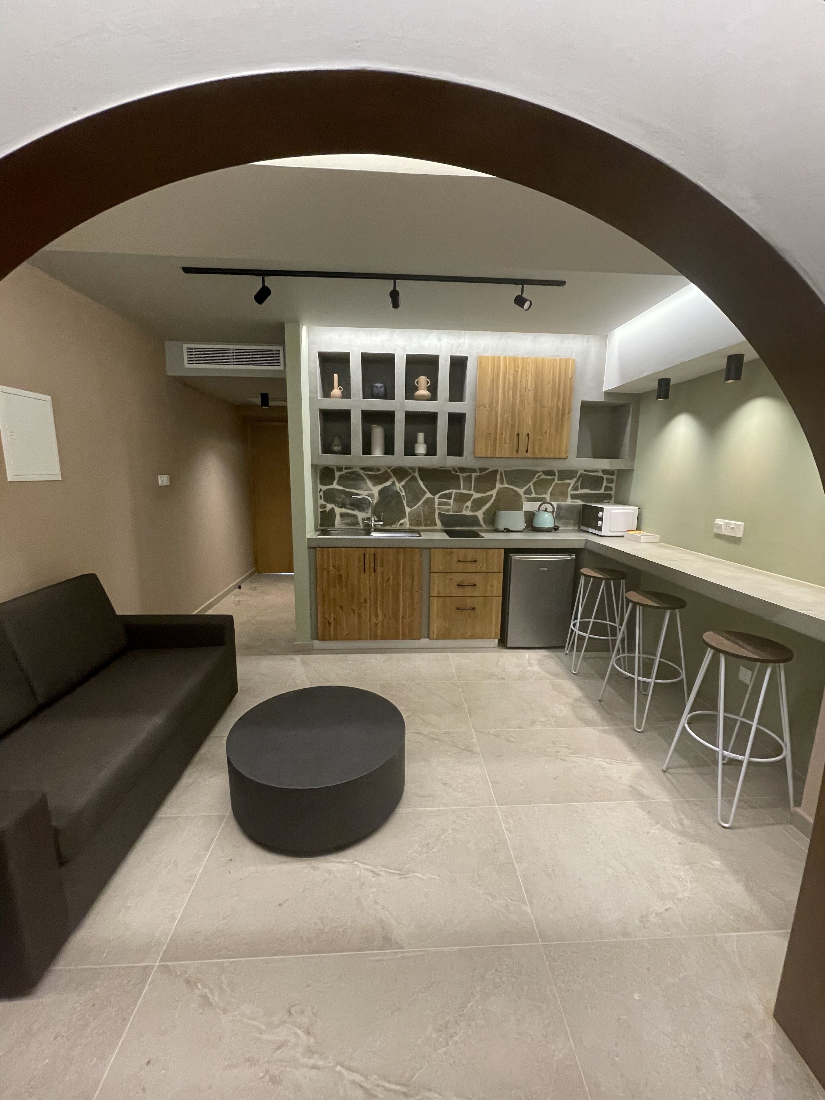 Een moderne keuken met een achterwand van steen, houten kastjes en een zwart poefje. Er is een bar met krukken en een ingebouwde oven.