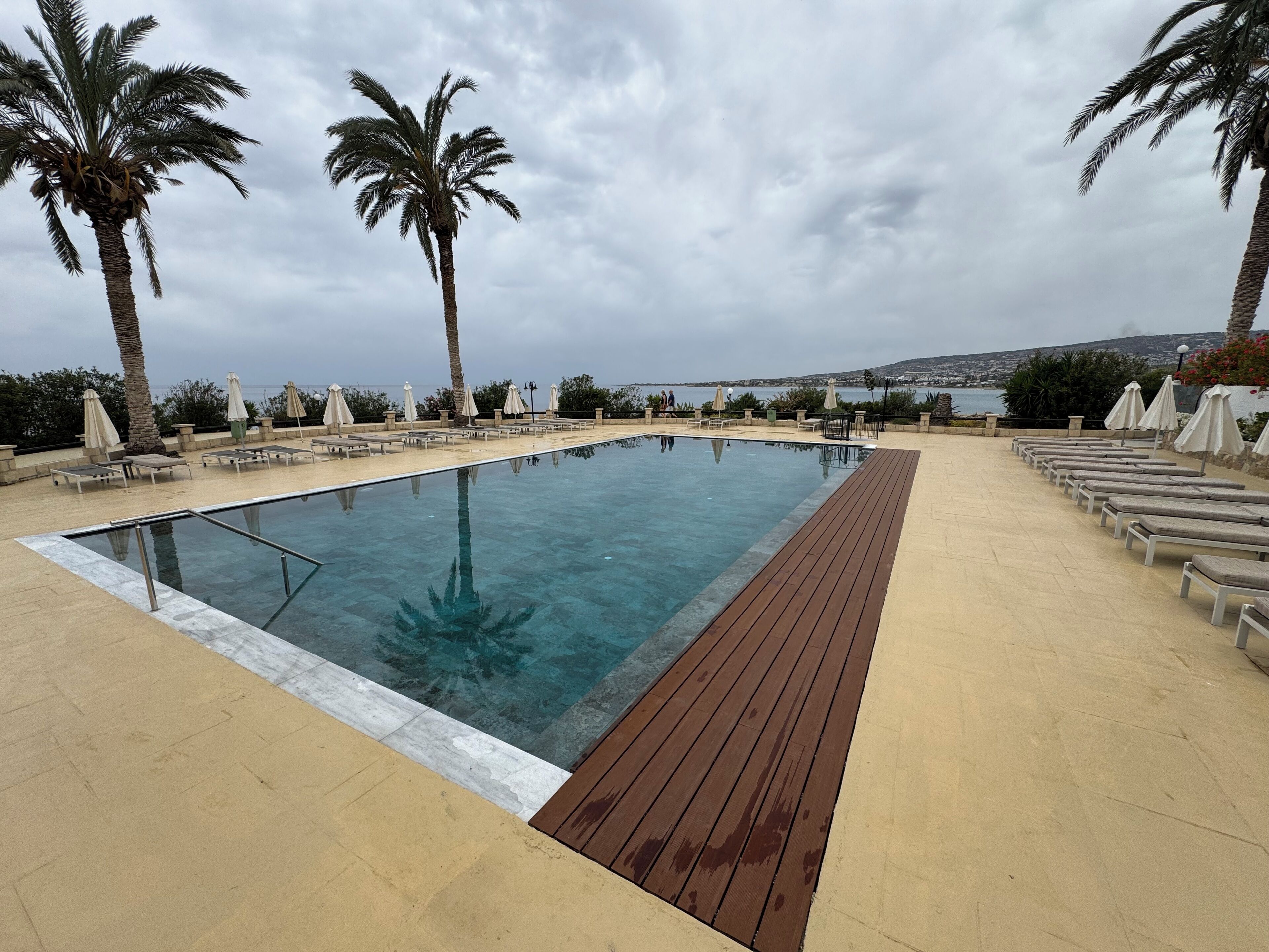 Une piscine rectangulaire dotée d’une terrasse en bois, entourée de chaises longues et de parasols, avec des palmiers et une vue sur la mer.