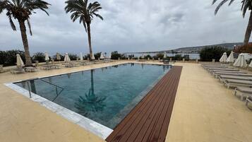 Une piscine rectangulaire dotée d’une terrasse en bois, entourée de chaises longues et de parasols, avec des palmiers et une vue sur la mer.