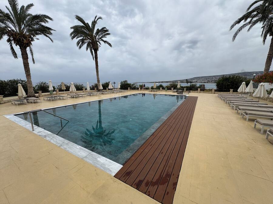 Une piscine rectangulaire dotée d’une terrasse en bois, entourée de chaises longues et de parasols, avec des palmiers et une vue sur la mer.