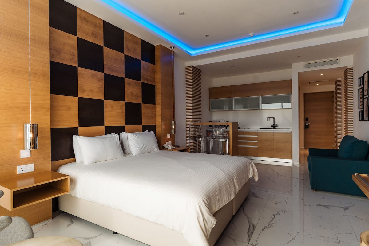 A modern hotel room with a large bed, a wooden headboard, a built-in desk, and a kitchenette.