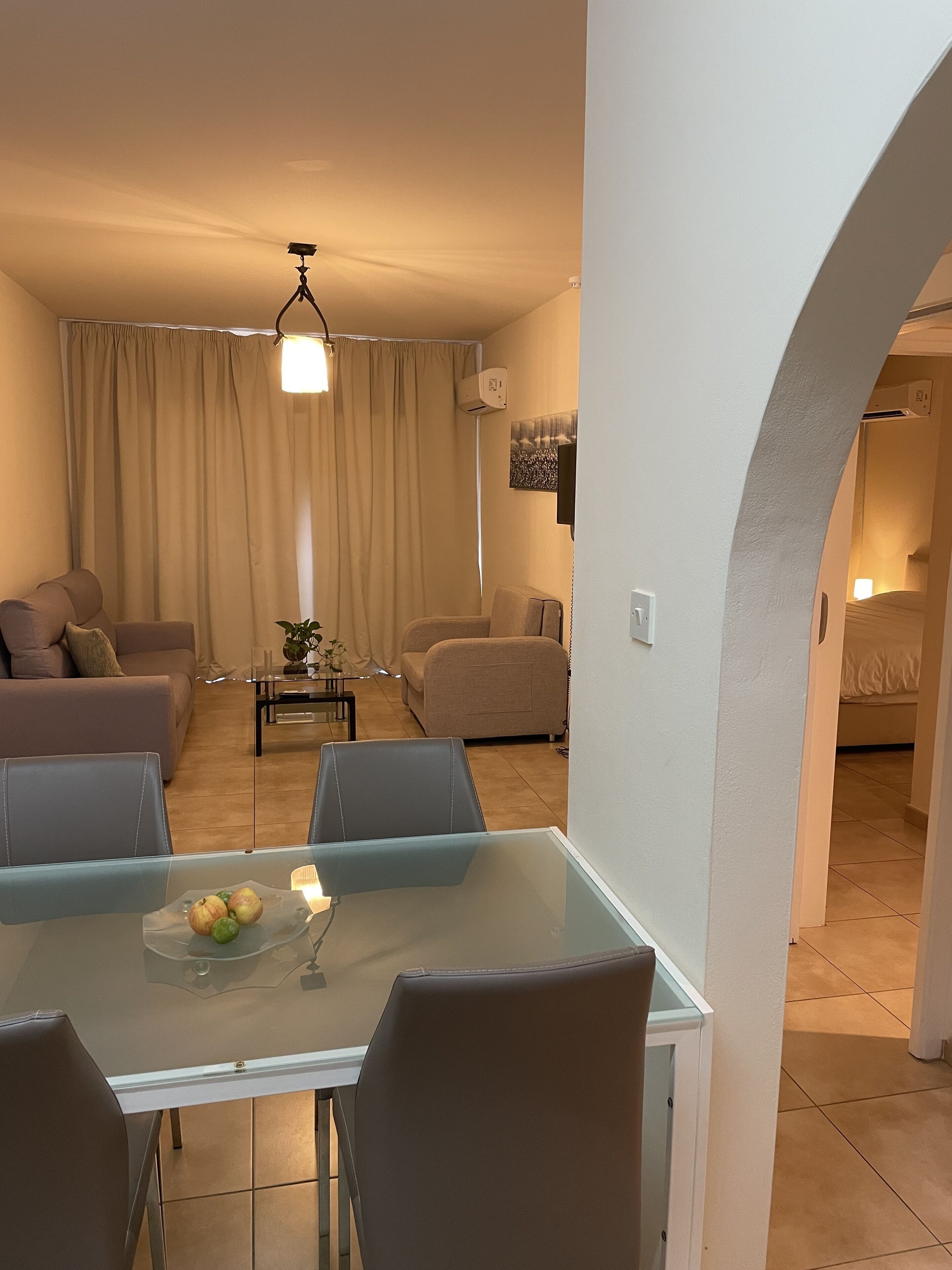 Una sala de estar moderna con una mesa de comedor de vidrio, dos sofás, un televisor montado en la pared y una lámpara de techo.